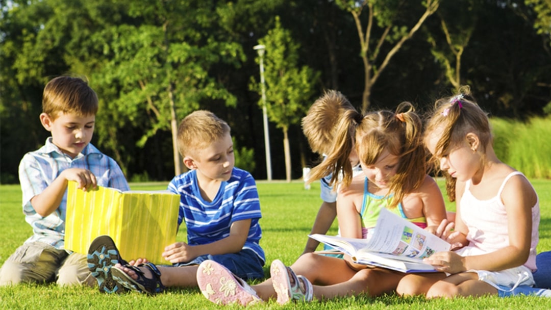 A group of young children reading together outside sat on grass