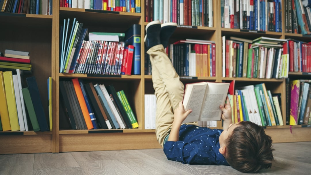 Boy reading a book on his back with his legs resting on a bookcase